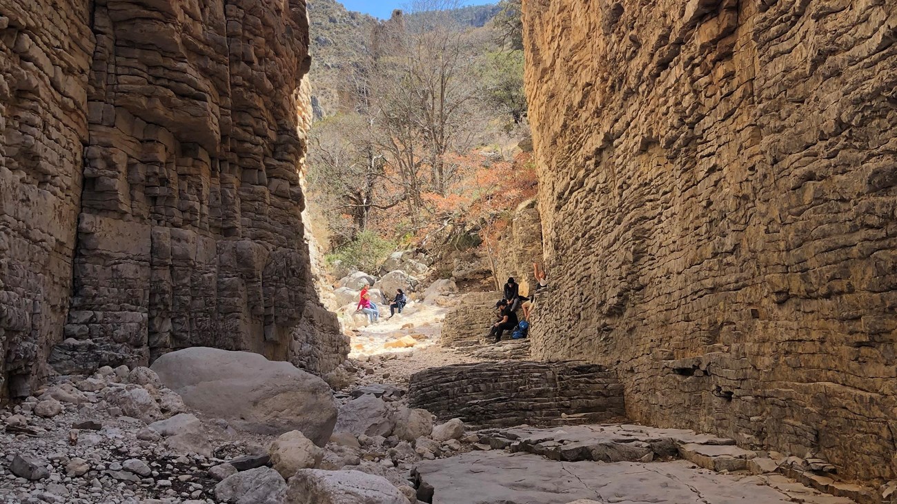 Groups of people in the sunlight at the end of a narrow tall canyon.