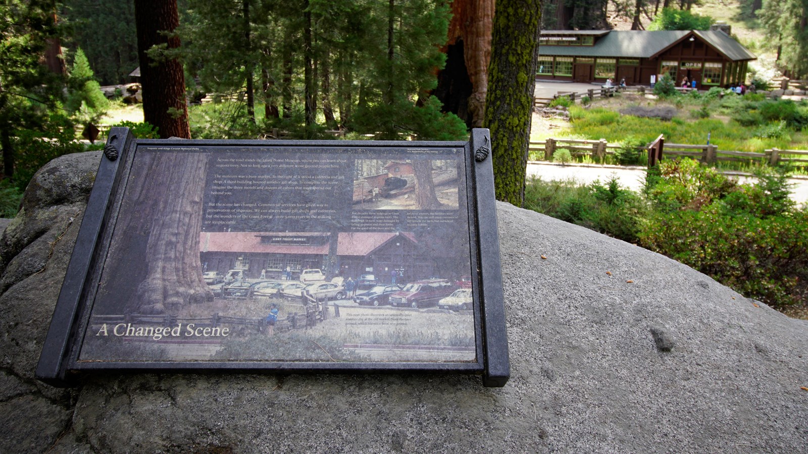 A black metal panel is bolted to a granite rock, in the distance behind it is a large brown building