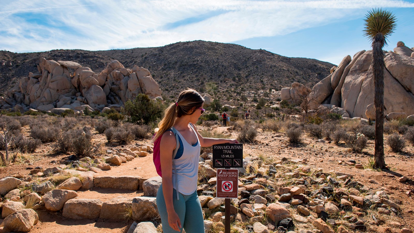A hiker next to a sign for Ryan Mountain by a dirt and rock trail heading towards a mountain.