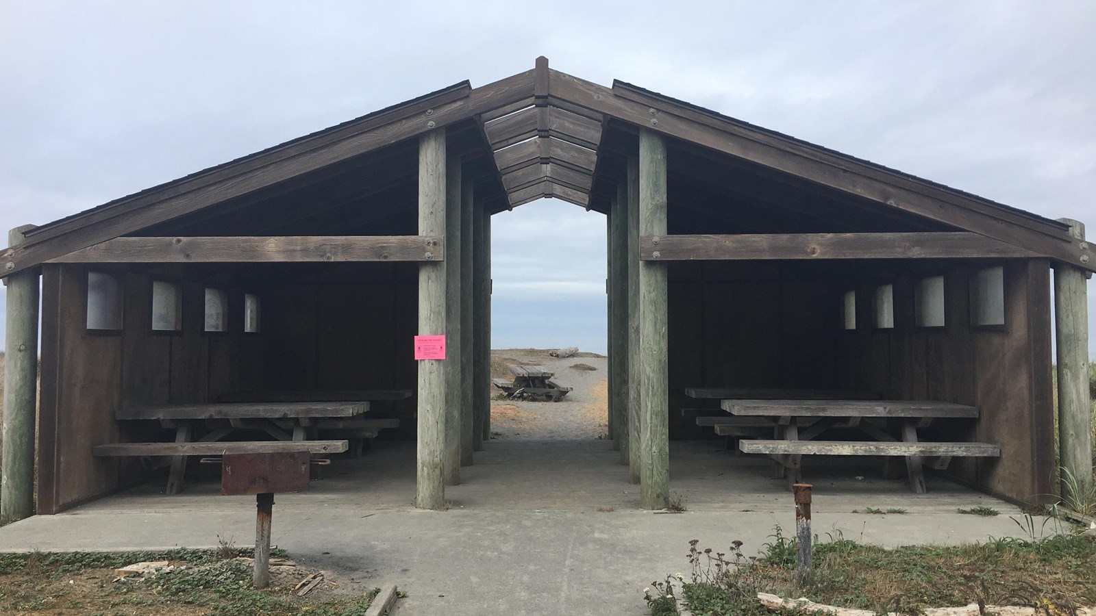 A wooden shelter protects picnic tables. A breezeway leads to a sandy beach.