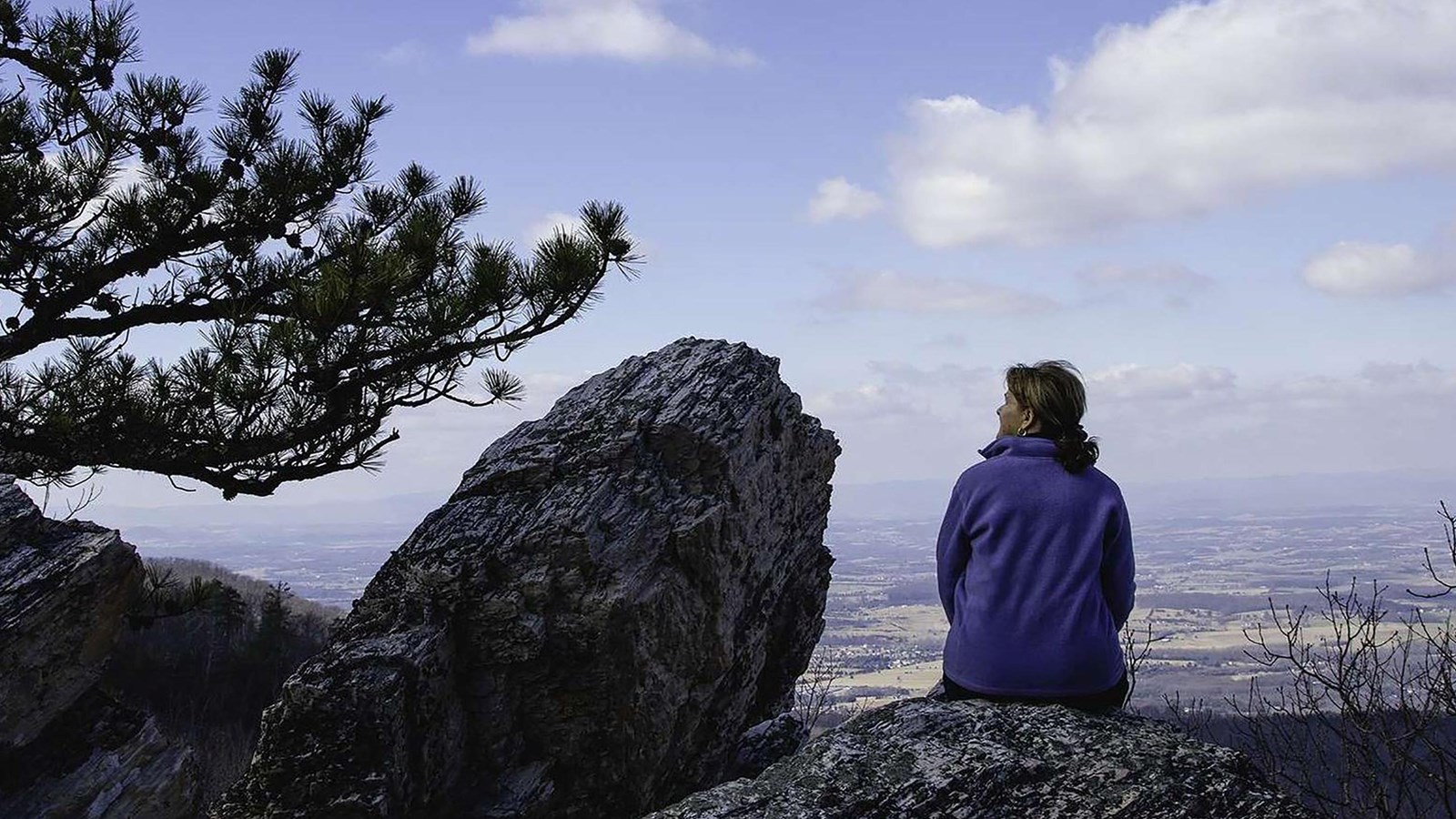 A color photograph of a woman sitting on a rock atop a mountain looking to the side.