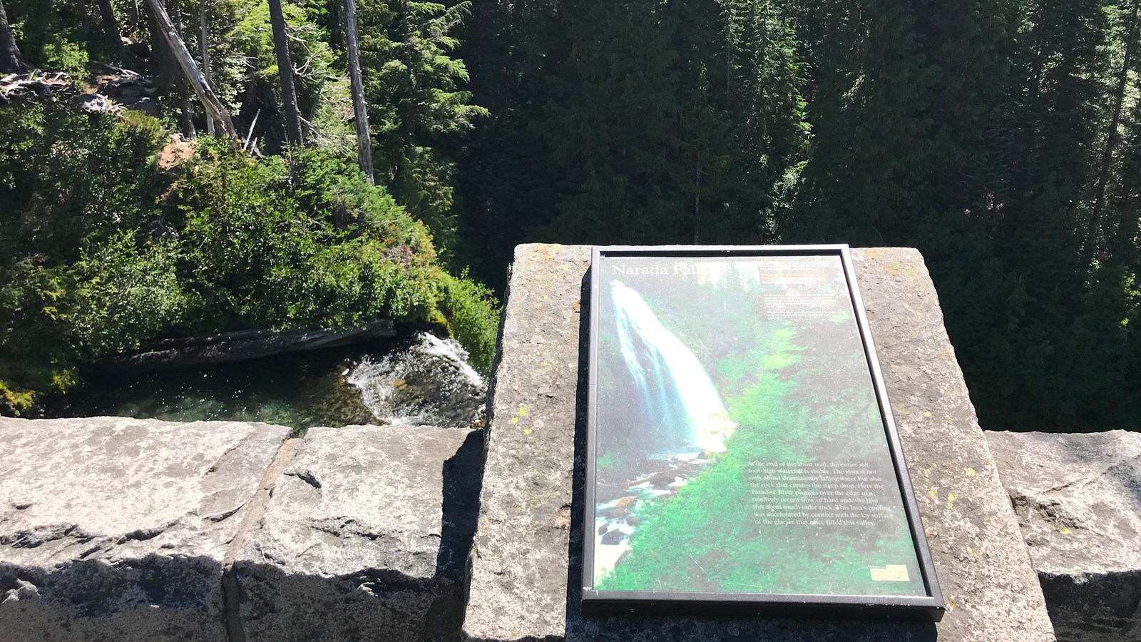A vertically-oriented wayside panel attached to a rock wall overlooking a river.