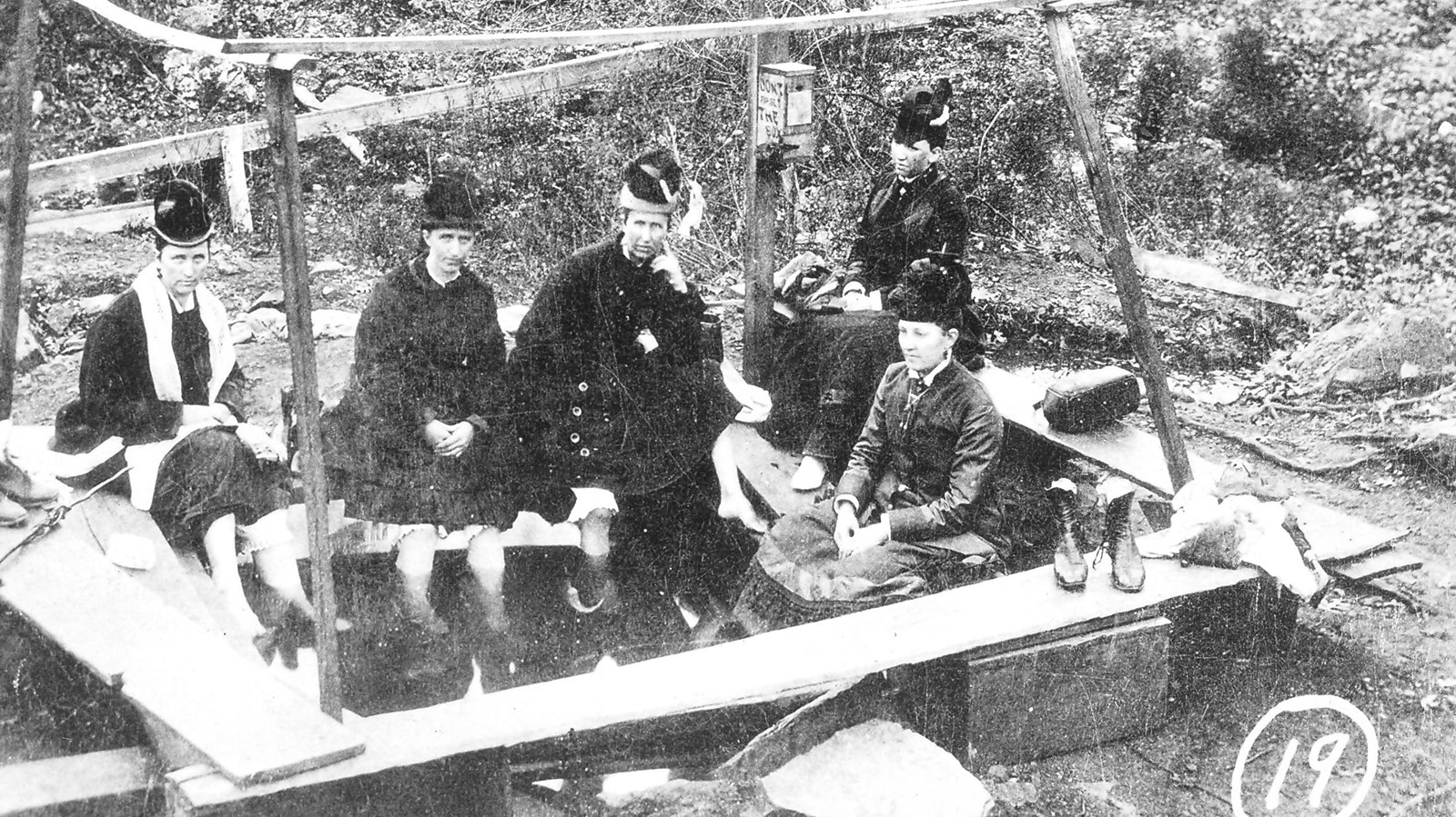 Five women in Victorian-style clothing sit on wooden benches with their feet in a pool of water.