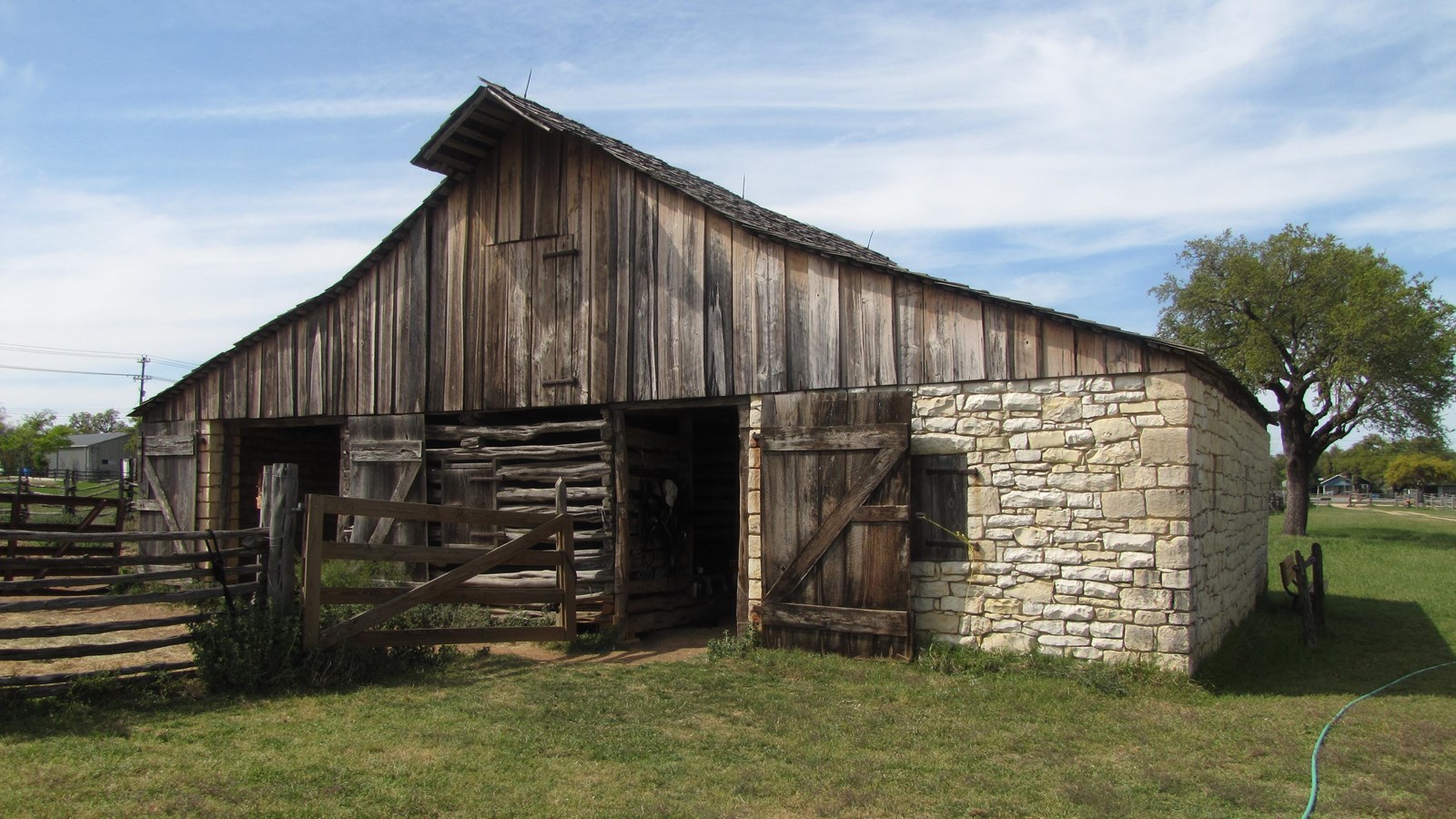 A barn constructed mostly of wood has two open doors, a fence and a large stone section.