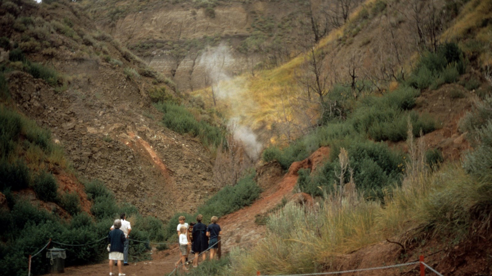 A small group of people stand at the base of a valley, watching smoke rise from the ground. 