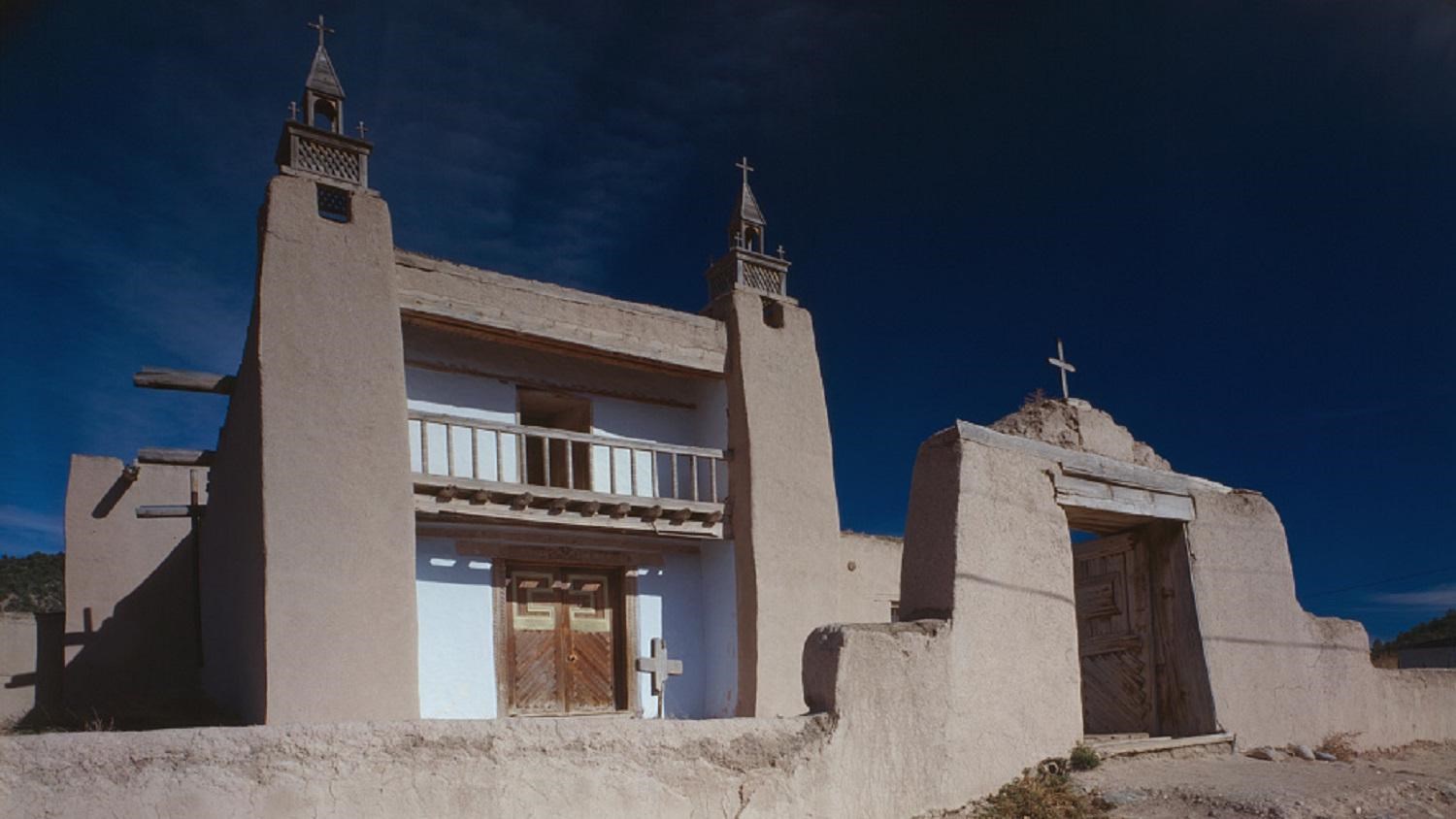 Two story adobe building with tan towers on either side of a white washed door and balcony.