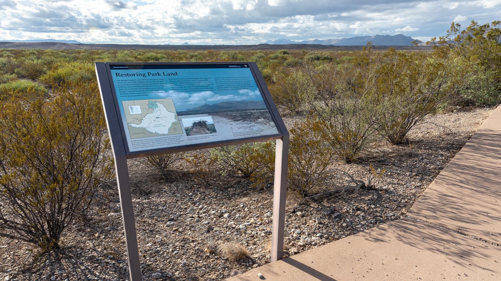 A metal sign is perched on the edge of a sidewalk overlooking the open desert with mountains behind.