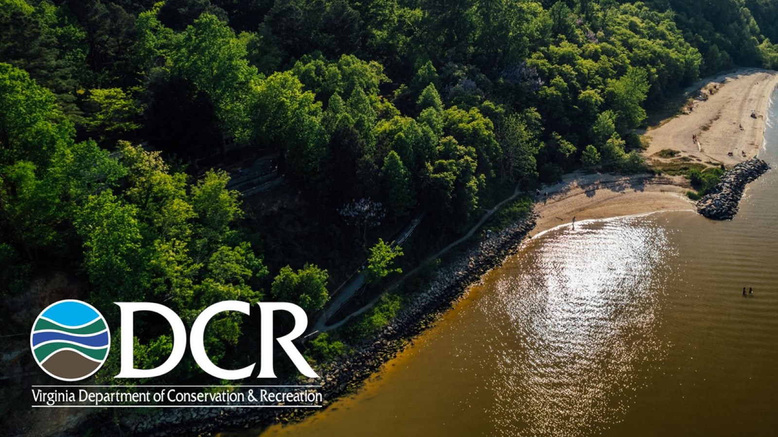 Aerial view of a forested shoreline with a sandy beach, rocky breakwaters, and people near sunlit wa