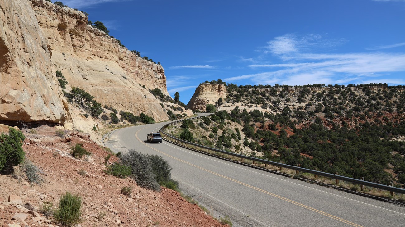 A card drives on a road that winds by yellow and red colored cliffs.