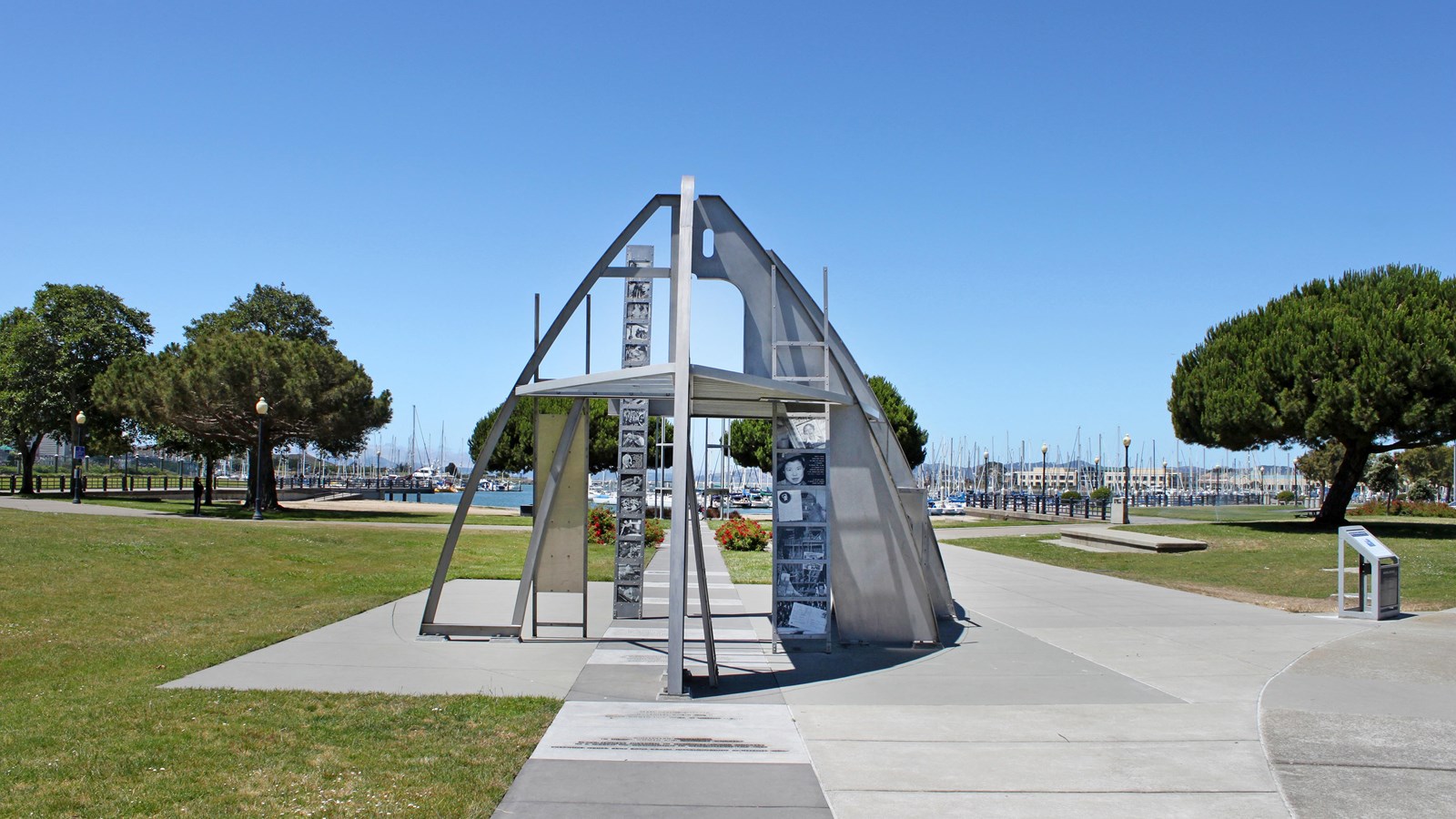 Metal structure, part of a ship build that contains photos. Sidewalk, trees and marina bay. 