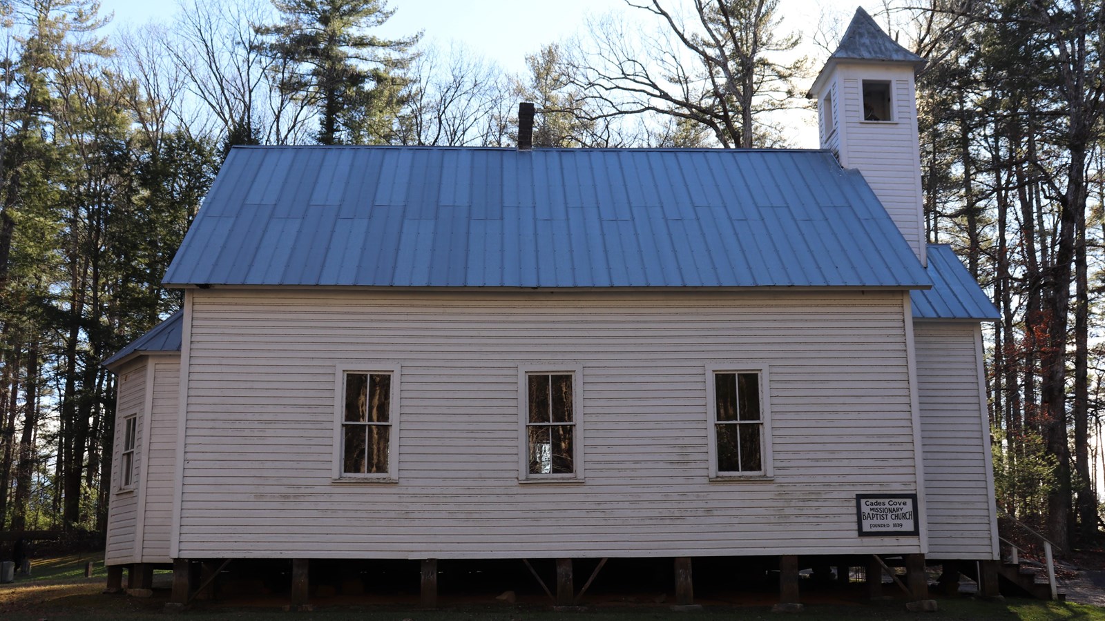 A white church with a blue roof in a field. Three rectangular windows and a sign are along the side.