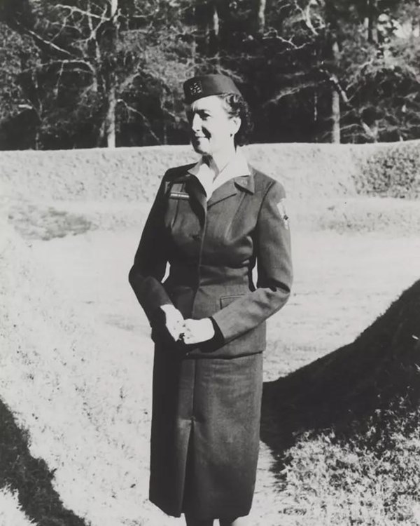Black and white photo of a woman wearing a skirt, jacket and pill box hat standing at the earthwork