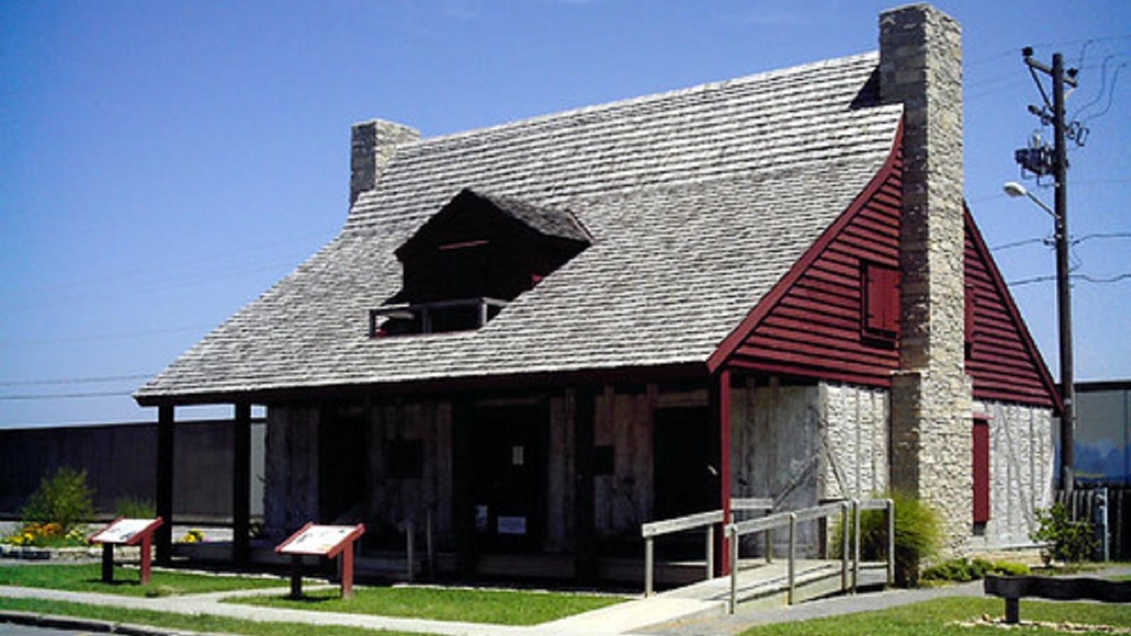 A white field stone and red-washed building with two chimneys sits along side a paved road.