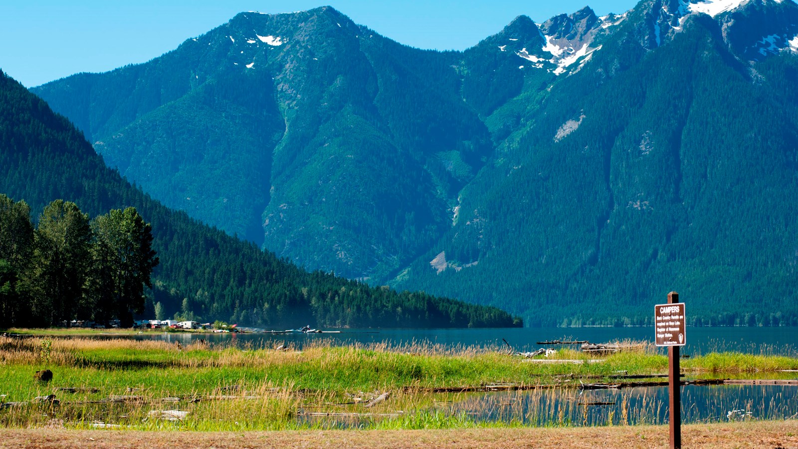 A sandy point in front of a large lake surrounded by mountains and forests.