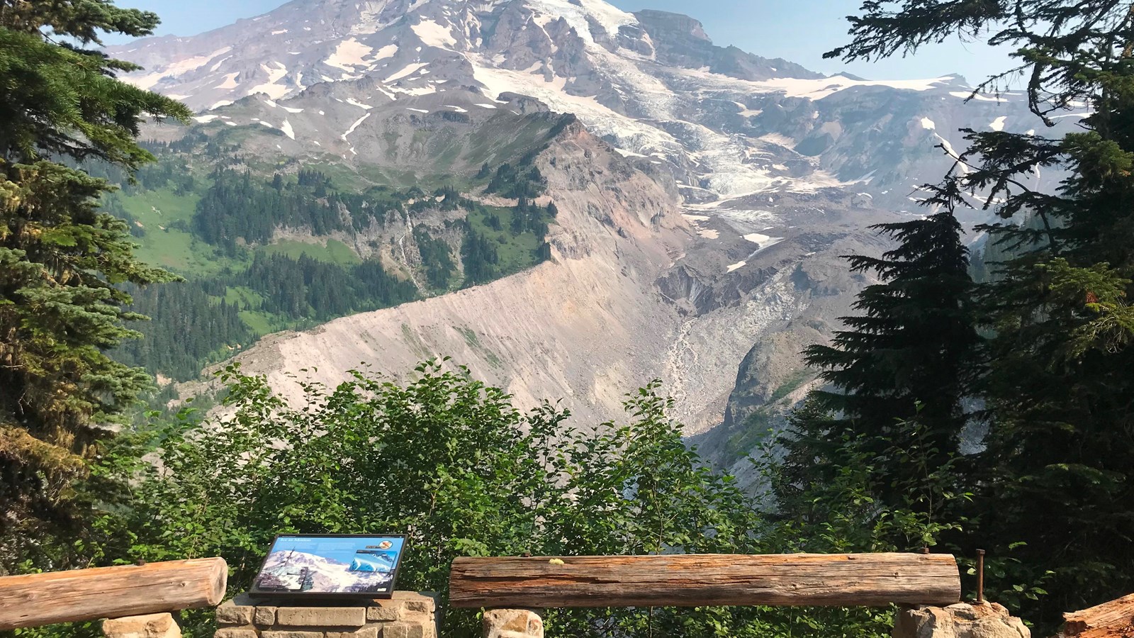 A wayside panel attached to a low rock and wood fence overlooking a glacier filled valley.