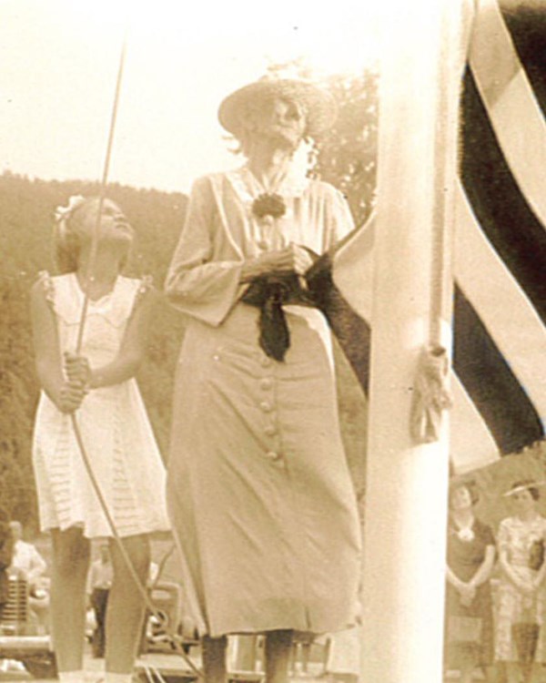Elderly woman in dress holds end of American flag as faded young girl in dress pulls rope on flag po