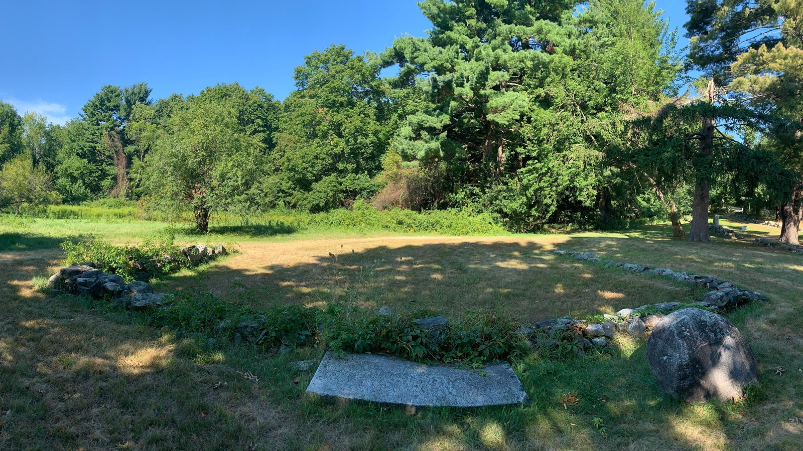 A low stone foundation rises from a meadow surrounded by trees. There is a large flat stone in front