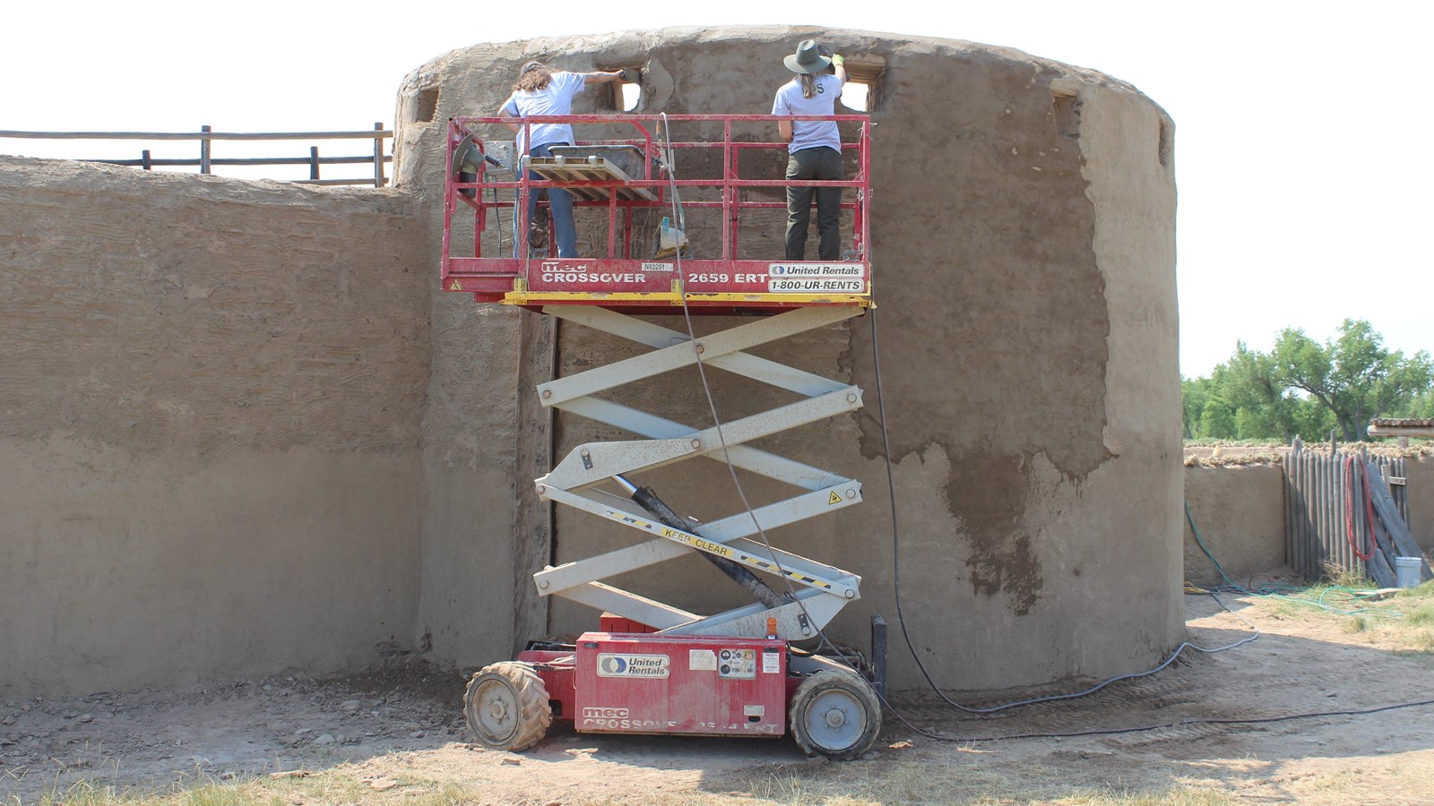 A scissor lift elevates two employees along a round wall to conduct repair work. 