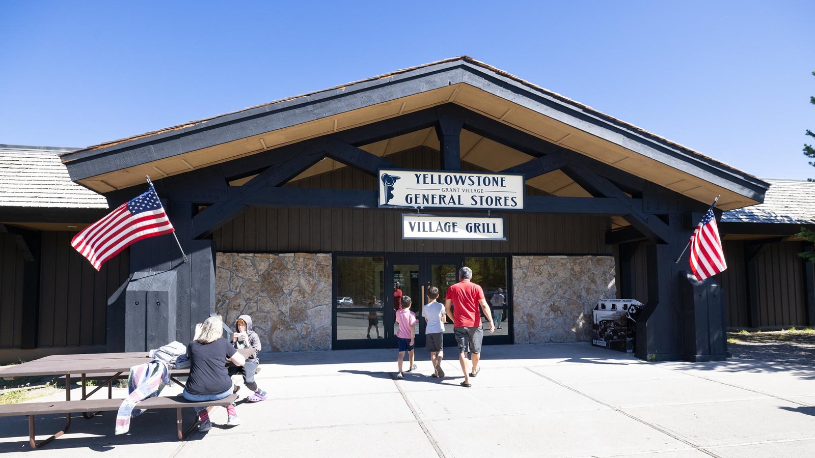 People walk towards a building with brown, vertical paneling and stone walls and an awning.