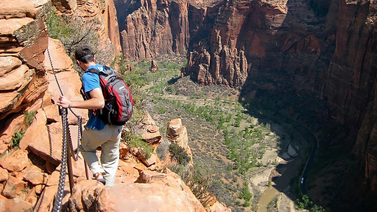 A hiker in a blue shirt holds onto chains as they traverse a sandstone cliff.