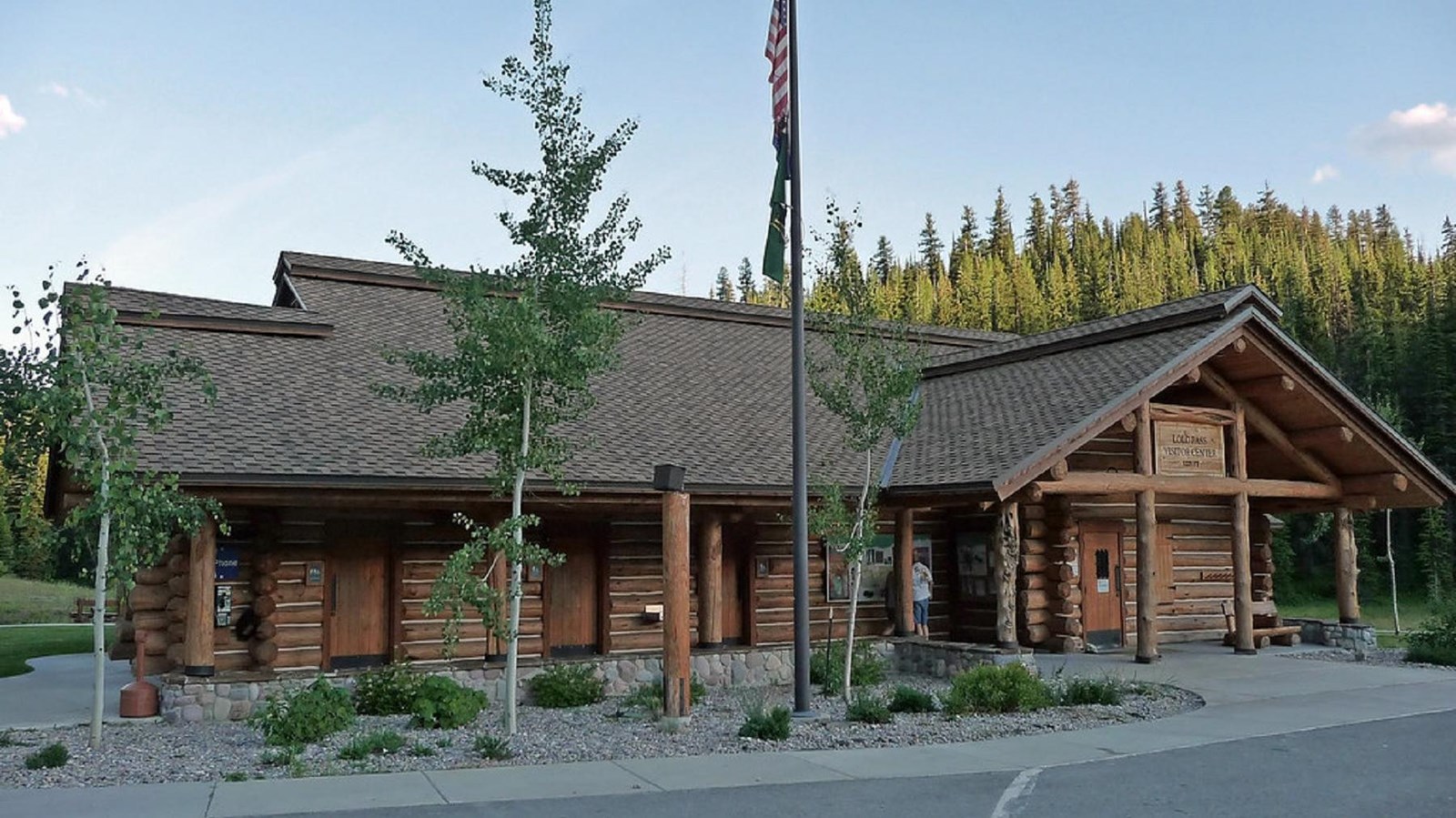 A rustic log building is flanked by deciduous trees and a flagpole