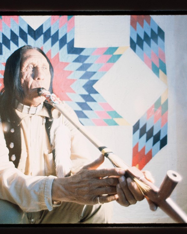 A seated in front of a quilt holding a pipe 