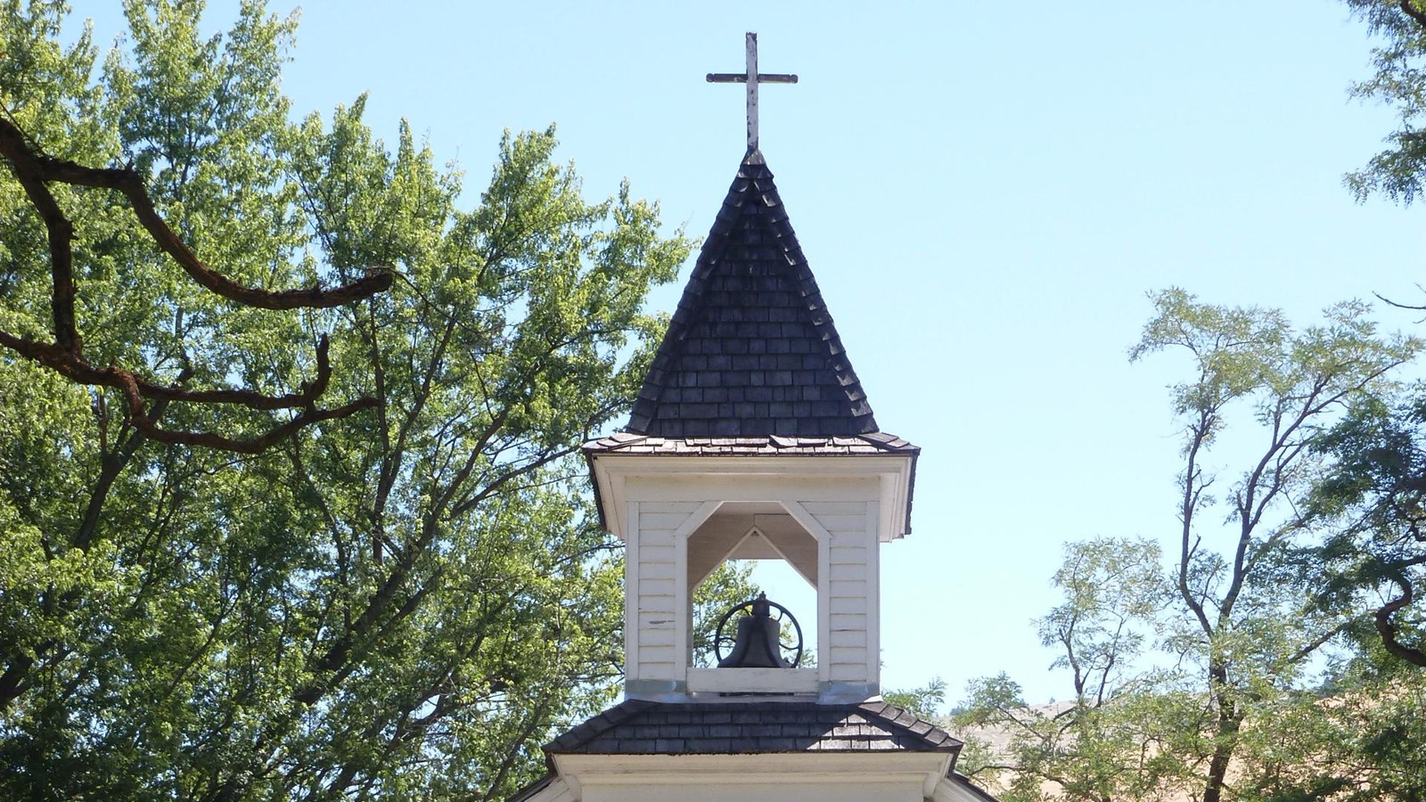 White mission with a church bell under black steeple roof with a cross on top. 