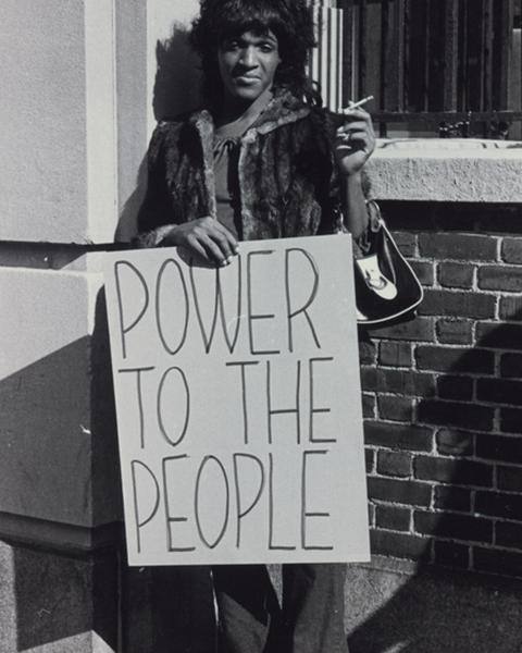 Marsha stands in front of a brick wall holding a sign that says Power to the People. by Diana Davies