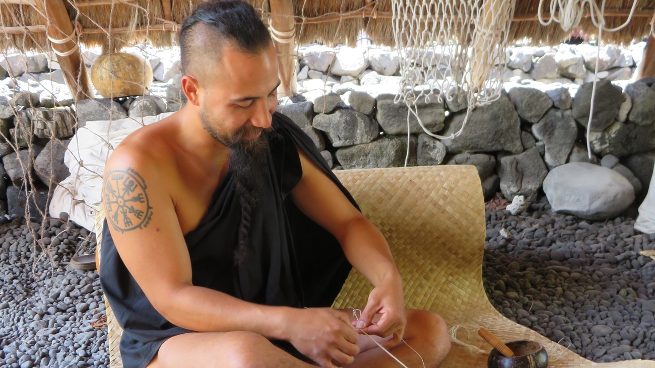 A man in traditional clothing weaves while sitting on a lauhala mat