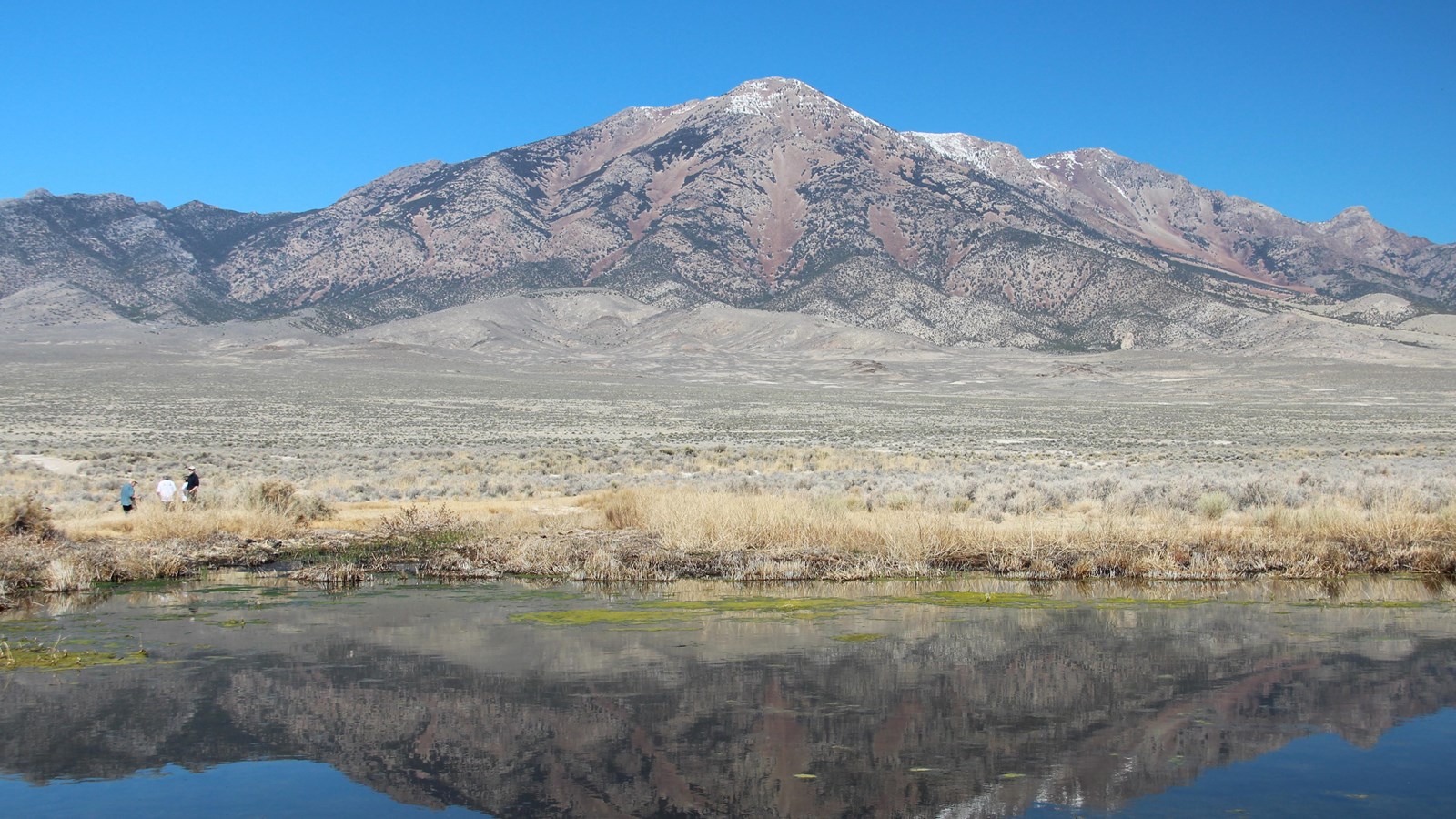 Clear spring pool reflects Pilot Peak; sagebrush plain and snowy ridges rise under a deep blue sky.