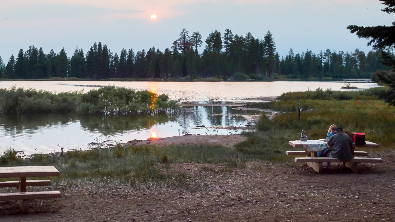 A couple sits at one of two picnic tables on the shore of a lake lined by conifers with the setting 