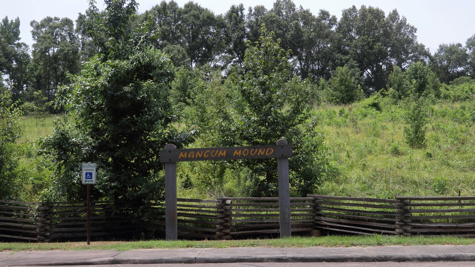 A hill of grass surrounded by trees, a split rail fence and wooden overhead sign, Mangum Mound