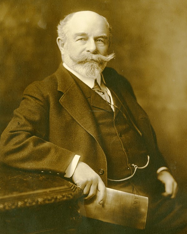 Studio portrait of man with large, shaped mustache in three-piece suit and tie, holding magazine.