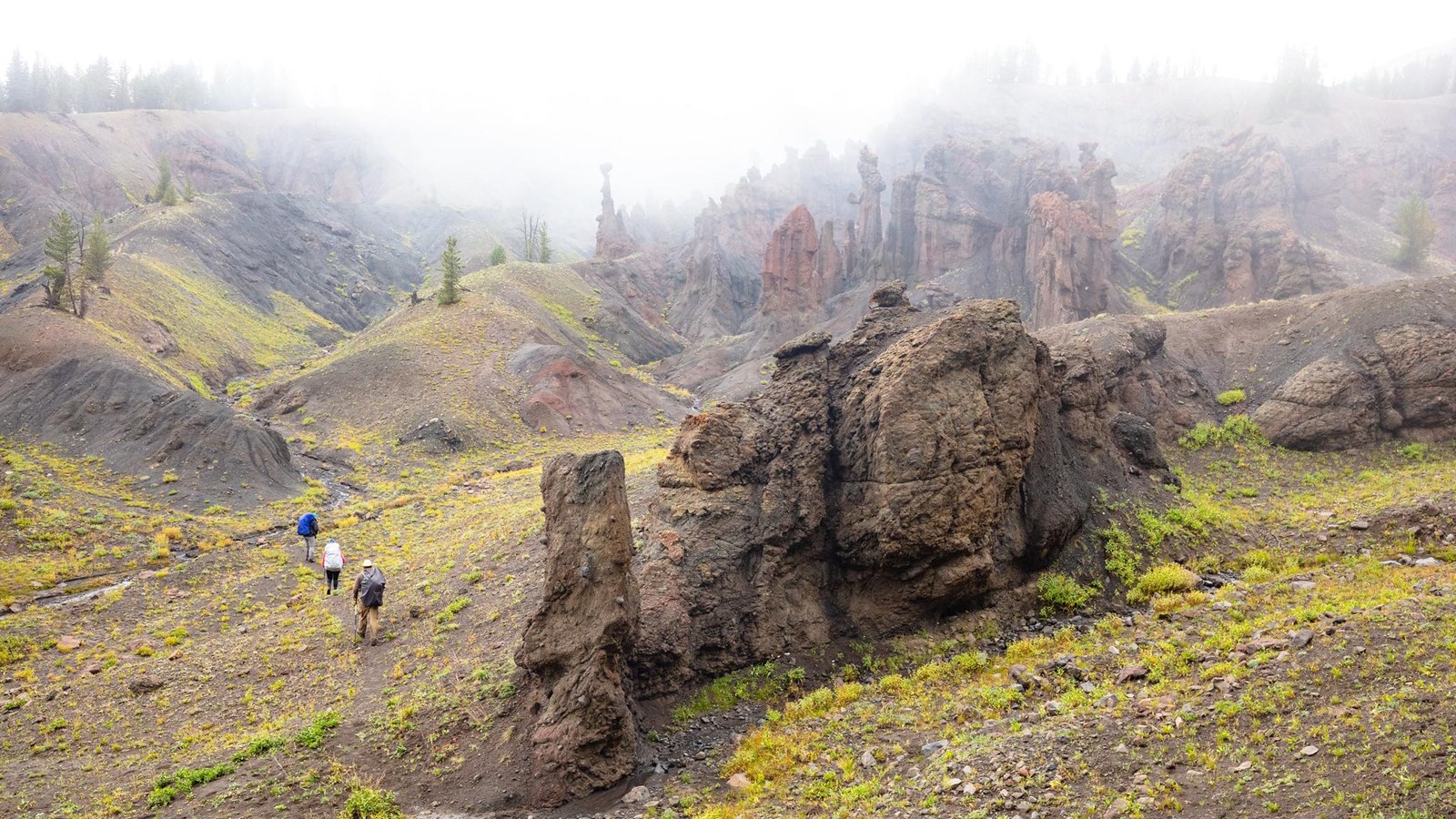 Three people on a trail hike up a drainage surrounded by rock formations on a rainy day.