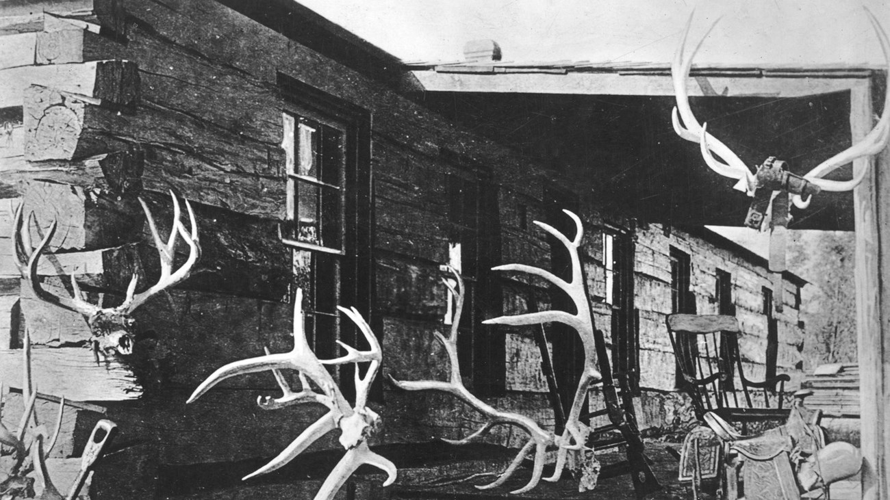 A black and white photo of a low ranch house, with a porch. Elk antlers hang on the walls.