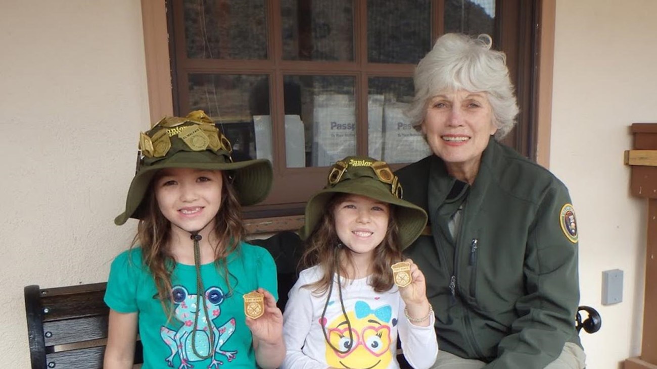 Two kids sit on a bench with a park volunteer. They\'re holding up badges.