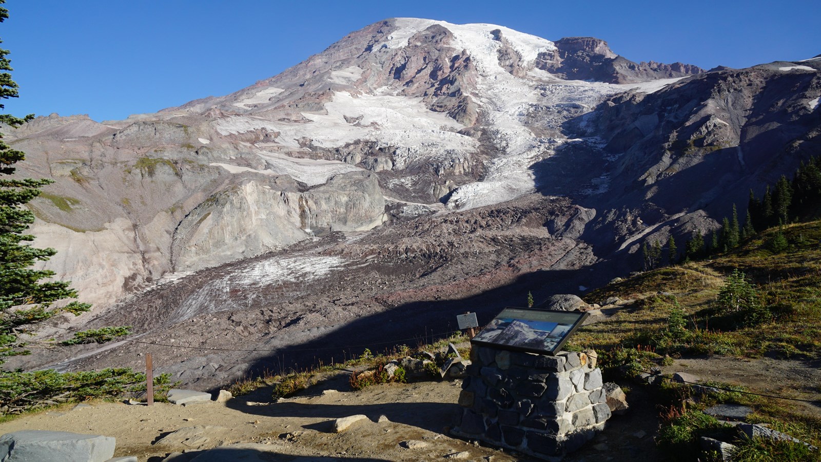 A wayside panel attached to a rock stand overlooking a glacier and mountain peak.