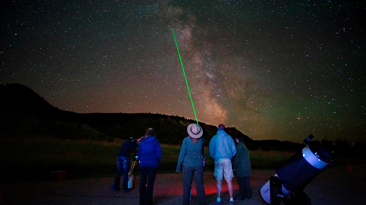 A ranger stands with several people, pointing a green laser at the sky. Stars are visible above them