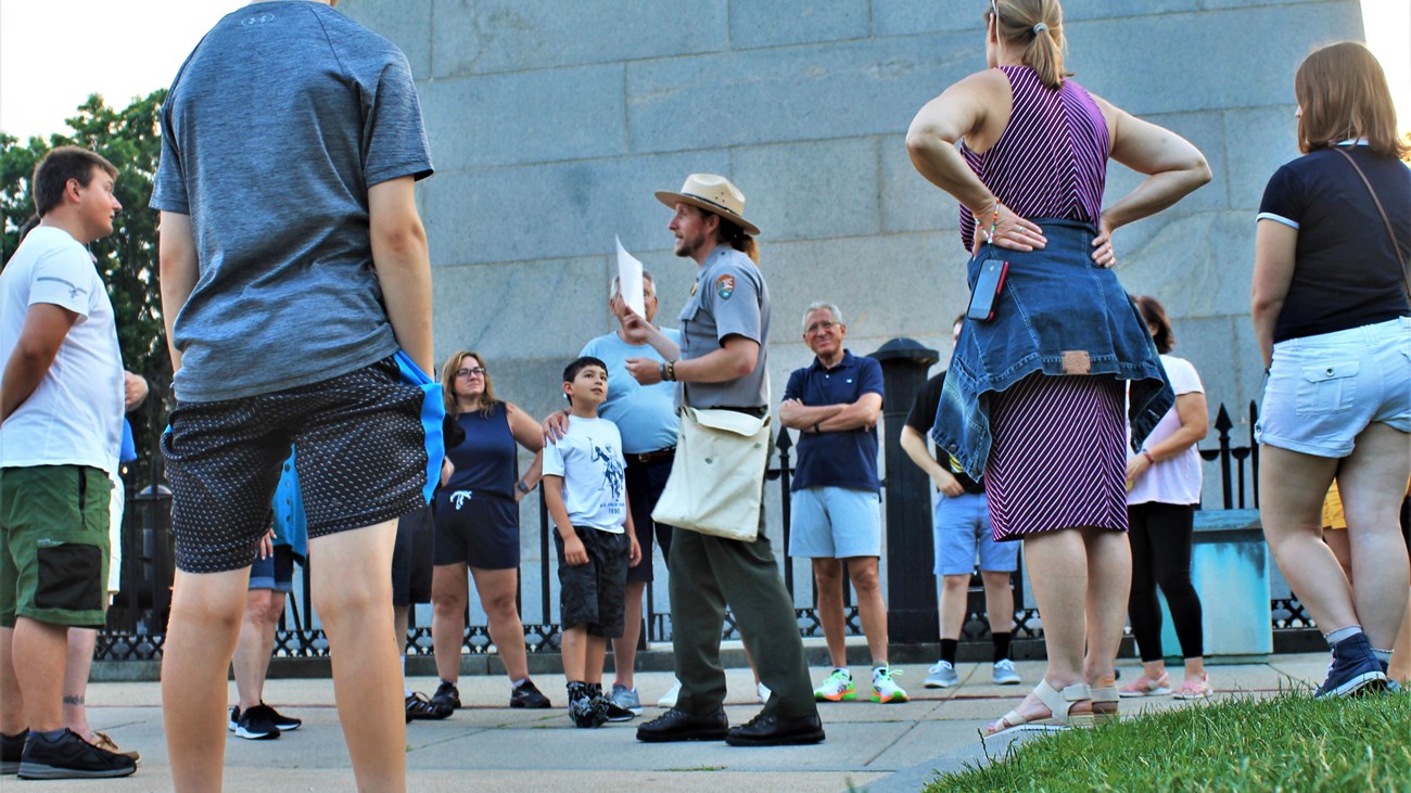 Ranger speaking in front of the Bunker Hill Monument to a group that surrounds him in a circle.