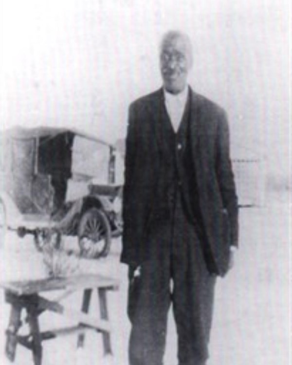 b&W photo, Black man in black suit with white button-down shirt stands outside