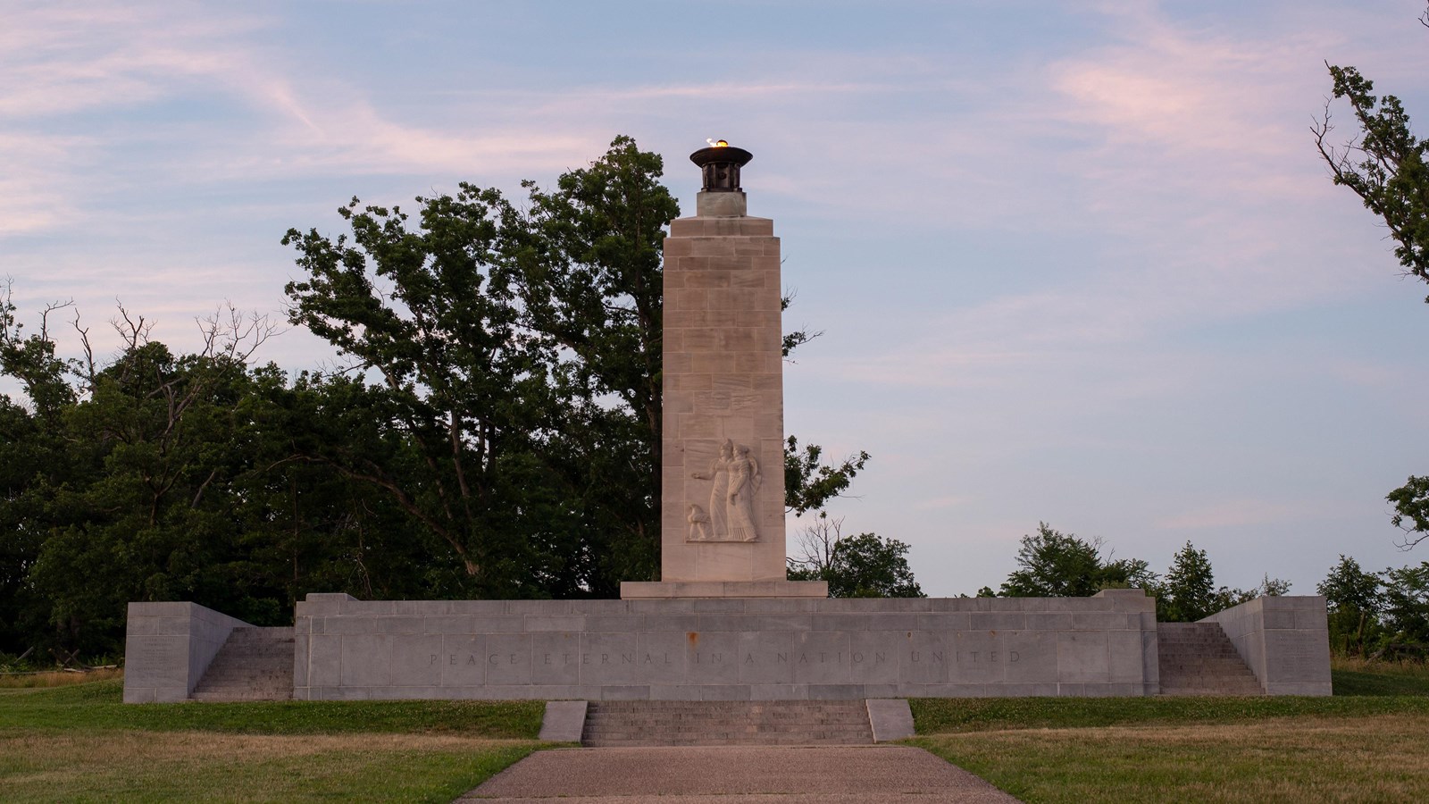 A white marble monument on a grey pedestal with storm clouds in the distance 