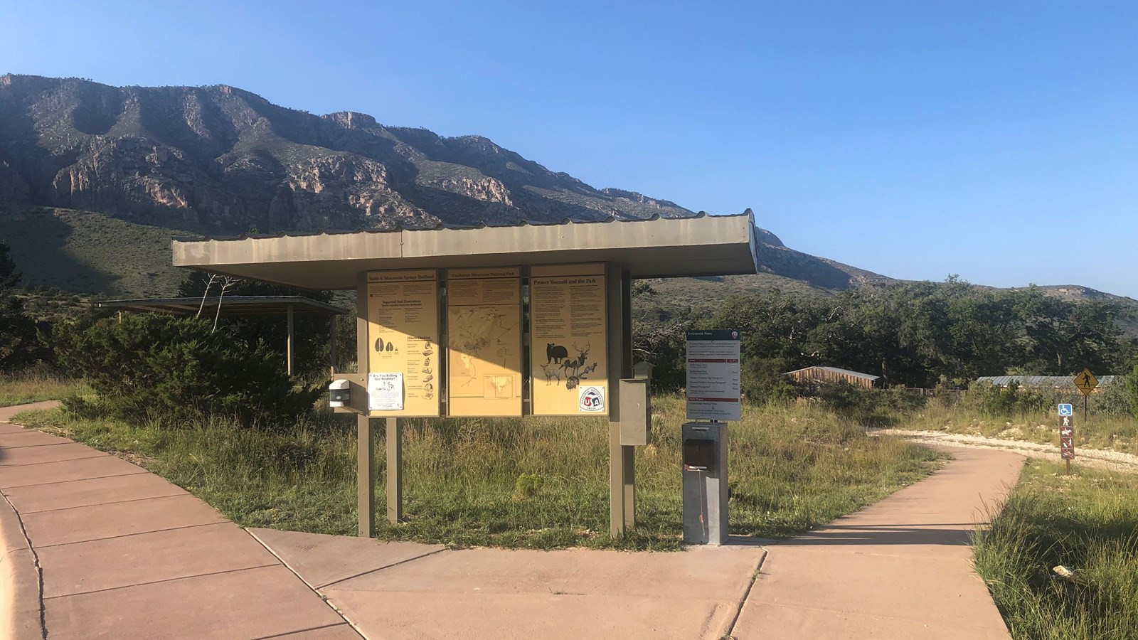 A metal kiosk with trail information stands before a historic ranch compound.