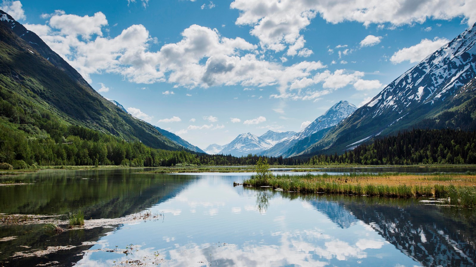 glassy lake reflecting puffy white clouds in the sky, tall mountains on either side