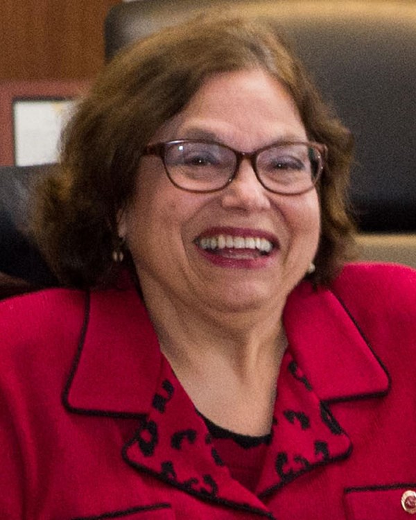 Judy, a woman with shoulder length brown hair smiles at the camera, she is wearing a red shirt.