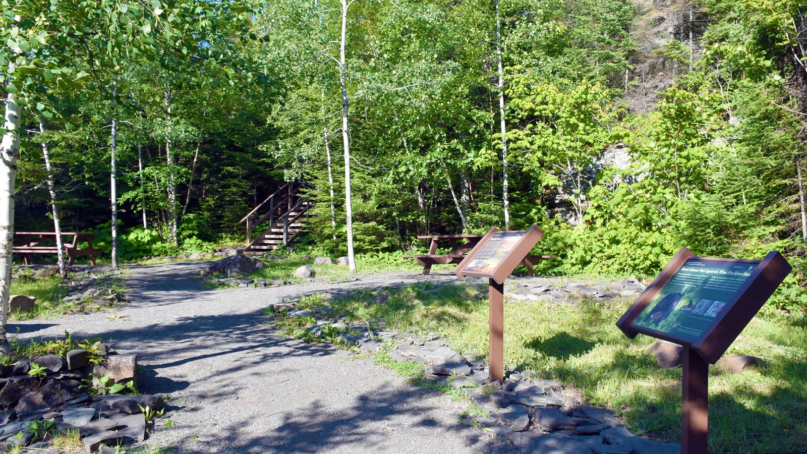 Gravel pathway between picnic tables to wooden stairs. Two interpretive signs in the foreground.