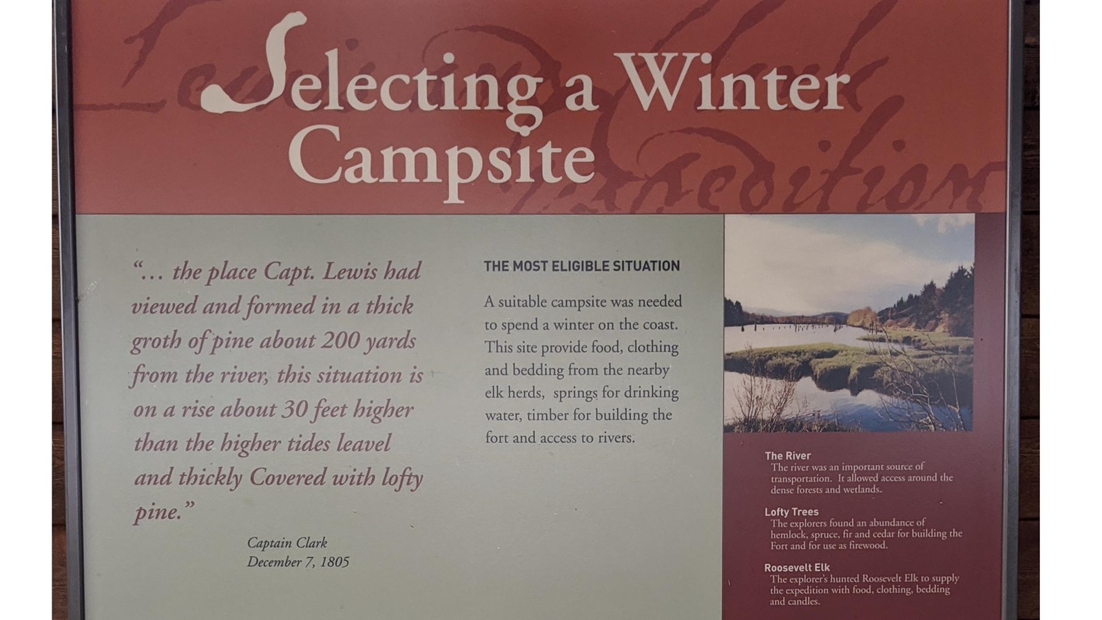 A sign hangs on wooden planks in an open air shelter. An elk, a river, and descriptions on it.