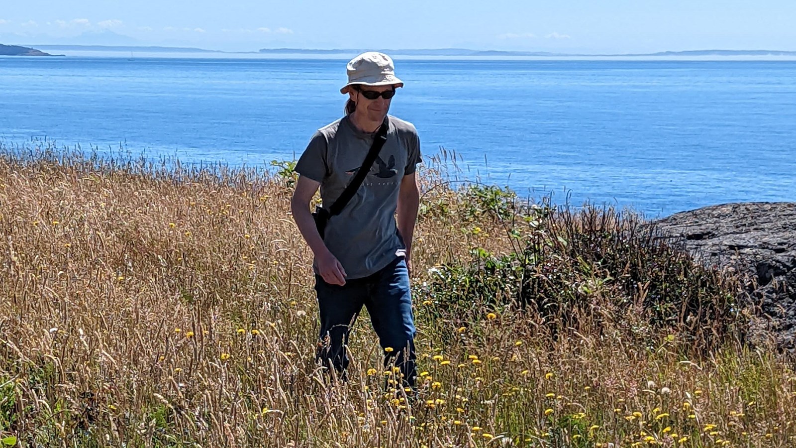 Color photo of a man hiking on a small trail that cuts through grassland. Behind him is the ocean