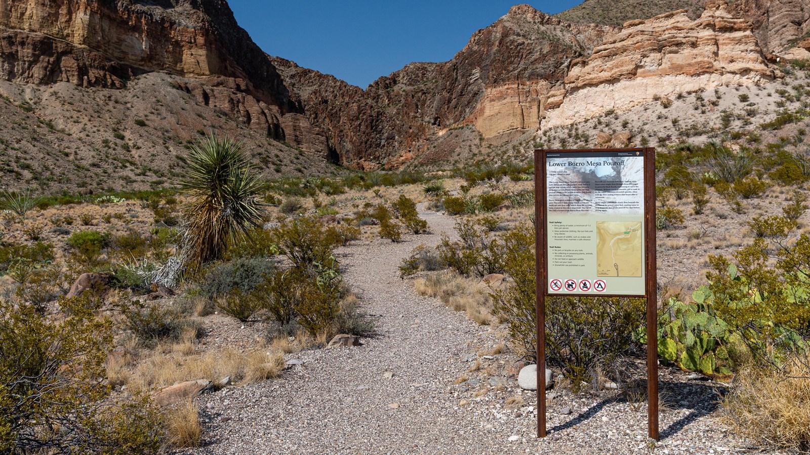 A gravel path starts at a trailhead sign and winds back to colorful igneous cliffs.