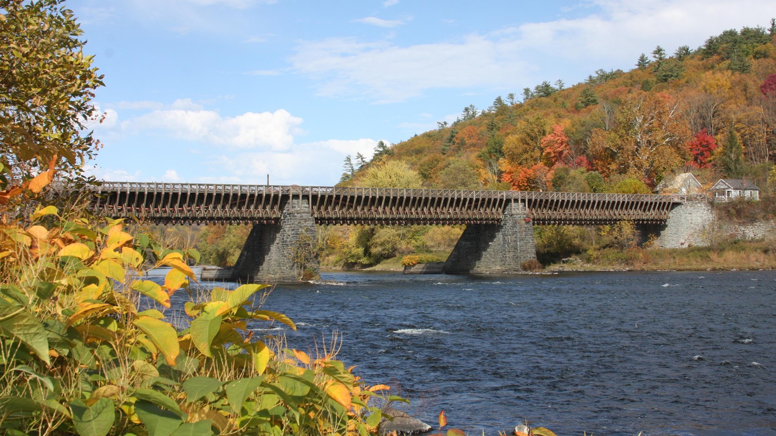 Wood bridge with two stone supports over a river. Trees in orange, green, and yellow color hills.