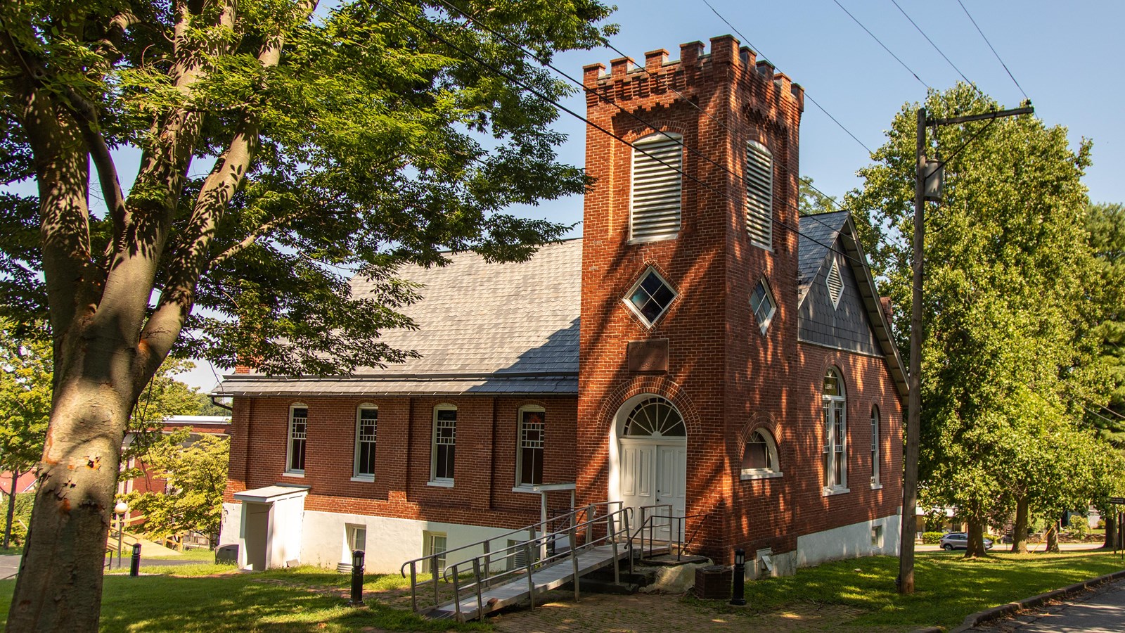 A brick church stands in the shade of a large tree next to the road.